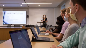 An instructor at the front of a classroom speakings while standing in front of a whiteboard and next to a screen as a row of students seated at a table with laptops listens.