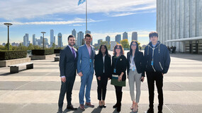 A group of students standing in a concrete plaza with the U.N. flag and the New York skyline behind them