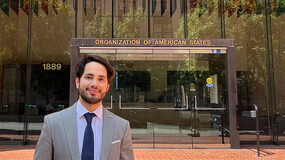 A man wearing suit stands before a glass-fronted building with the words "ORGANIZATION OF AMERICAN STATES" and a row of flags from various countries above the door.
