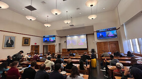 An audience seated in a classroom watching to panelists in the room and on screen