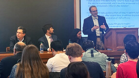 A speaker at a podium with the words "FDA and the Public Trust: Ca0se 1" projected onto a screen while three panelists are seated in front of a chalkboard and audience members sit in rows