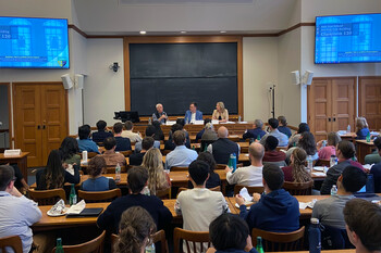 students in a crowded classroom listening to three speakers seated at the front of the room